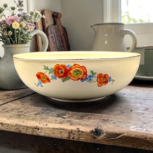 May include: A cream-colored ceramic bowl featuring a decorative floral pattern of vibrant orange poppies and small blue flowers along the exterior side. The bowl has a smooth, rounded shape with a thin metallic rim detail around the top edge.