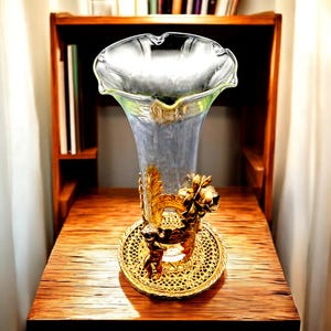 May include: A clear glass vase with a flared rim and a gold-colored base. The base features ornate detailing, including a cherub and floral accents. The vase is displayed on a wooden surface, with a bookshelf in the background.