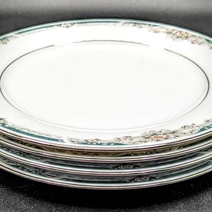 May include: A stack of four white ceramic plates with a decorative floral and teal border. The plates have a gold trim and are stacked on top of each other. The floral design includes pink and beige flowers.