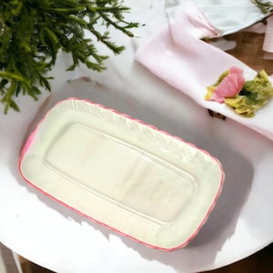 May include: A white rectangular serving dish with a pink rim. The dish is made of ceramic and has a slightly textured surface.