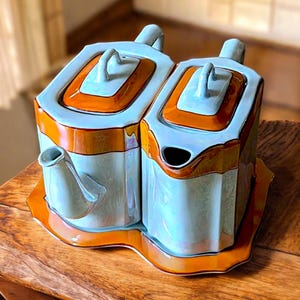 May include: A light blue and orange ceramic tea set with a teapot and creamer. The set has a geometric design with a light blue body and orange accents. The teapot has a spout and handle, and the creamer has a handle and open top. The set sits on a matching tray.
