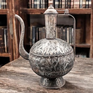 May include: A silver metal teapot with a rounded body and a long, curved spout. The teapot has a hammered finish and a decorative band around the middle. It is sitting on a wooden table.