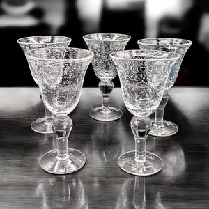 May include: Five clear, stemmed wine glasses with a bubble design. The glasses have a rounded bowl, a bulbous stem, and a circular base. They are arranged on a dark wooden surface, with a blurred background.