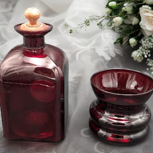 May include: A set of two dark red glass bottles with a wooden stopper on one bottle. The bottles are on a white and gray surface.