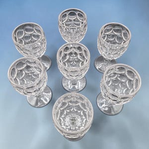 May include: Eight clear, stemmed glasses arranged in a circular pattern on a blue surface. The glasses have a textured, bubble-like pattern on the bowl and a simple, round base. The glasses are empty and appear to be made of glass.