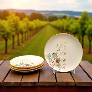 May include: Set of three cream-colored plates with a gold rim and a delicate botanical design featuring green, brown, and yellow leaves. One plate is displayed on a clear stand, while the others are stacked on a wooden surface.