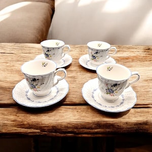 May include: Four white teacups and saucers with floral designs. Each cup and saucer has a blue trim and a delicate pattern of blue and pink flowers. The cups have curved handles and sit on a wooden surface.