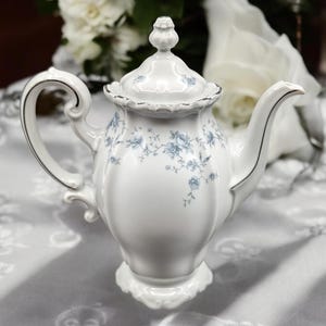 May include: A white porcelain teapot featuring a delicate blue floral pattern, a scalloped lid with a finial, a curved handle, and a slender spout. The teapot is accented with silver trim along the edges of the lid, handle, and spout.