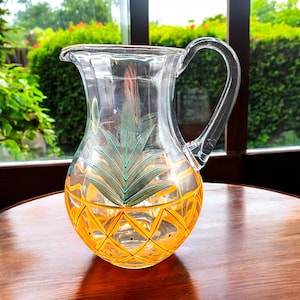 May include: A clear glass pitcher with a pineapple design. The pitcher has a yellow and gold pineapple pattern with green leaves.