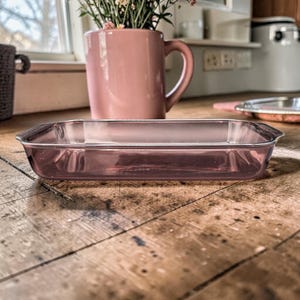May include: A rectangular baking dish made of translucent, light purple glass with integrated handles on both ends. The dish features smooth, rounded corners and a clear, polished finish, showcasing a simple, functional design for kitchen use.