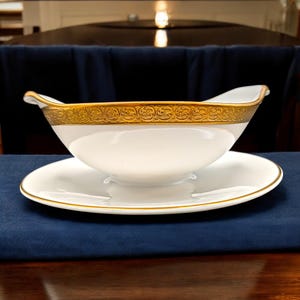 May include: A white porcelain gravy boat with a matching saucer. The boat has a gold ornate border and a gold trim on the rim and handle. The saucer also has a gold trim. The set is on a dark blue surface.