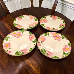 May include: A set of four round ceramic plates featuring a cream-colored base with hand-painted pink wild rose blossoms and green leaves along the scalloped edges. Each plate displays a consistent floral pattern with yellow centers on the flowers.
