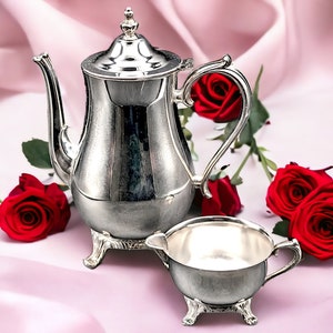 May include: A silver teapot and creamer set with a classic design. The teapot has a rounded body with a decorative handle and spout. The creamer is a smaller version of the teapot with a handle on one side. Both pieces are sitting on a pink satin surface with red roses.