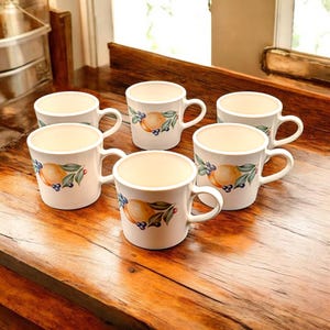 May include: Six white ceramic mugs with a cream-colored interior are arranged on a wooden surface. Each mug features a painted design of a yellow fruit, green leaves, and blue berries. The mugs have a classic handle design.