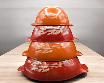 May include: A stack of four vintage Pyrex bowls in shades of orange and red. Each bowl features a wheat stalk design in white. The bowls are nested on a light-colored wooden surface.