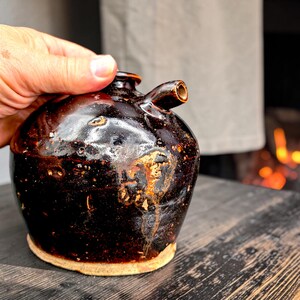 May include: A dark brown, glazed ceramic oil cruet with a rounded body, a small spout, and a narrow neck. The surface features a glossy, mottled finish with lighter tan patches and a visible, unglazed light beige base at the bottom.