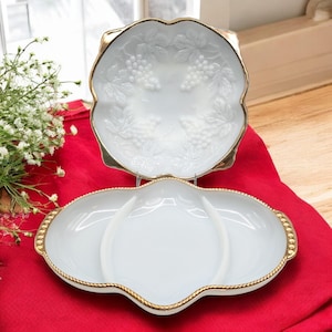 May include: Two white serving dishes with gold trim. The top dish has a scalloped edge and a grape design. The bottom dish is divided into three sections and has a beaded gold trim. Both dishes are on a red surface.