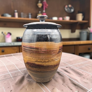 May include: A ceramic jar with a lid featuring a glossy black top section and a textured, multi-toned body with horizontal bands of earthy brown, ochre, and deep red glazes. The jar has a rounded silhouette and a small, knob-style handle.