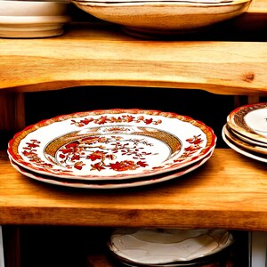 May include: A stack of white ceramic plates with a red floral design and gold trim. The plates are stacked on a wooden shelf with other dishware. The plates have a scalloped edge.