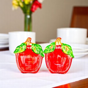 May include: Two red ceramic apple salt and pepper shakers with green leaves. The shakers are on a white tablecloth.