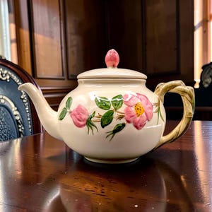 May include: A cream-colored ceramic teapot featuring a hand-painted floral design of pink wild roses with green leaves and stems. The teapot includes a matching lid with a pink bud-shaped finial and a handle textured to resemble a wooden branch.