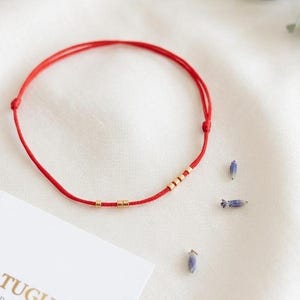 Könnte beinhalten: Ein dünnes rotes Schnurarmband mit fünf kleinen goldfarbenen zylindrischen Perlen, die in zwei Gruppen angeordnet sind. Das Armband verfügt über zwei verstellbare Schiebeknoten an den Seiten. Eine weiße rechteckige Karte mit dem Text RODICA TUGUI ist sichtbar.