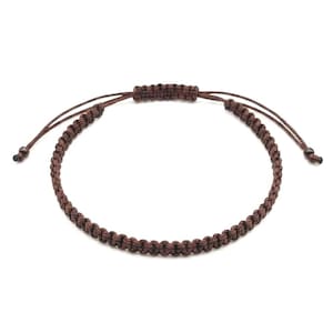 Op de afbeelding: Een donkerbruine koordarmband met een gevlochten patroon en een verstelbare schuifknoopsluiting. De armband is gemaakt van dun, geweven touw en bevat twee kleine kraaltjes aan de uiteinden van de trekkoorden voor een goede pasvorm.
