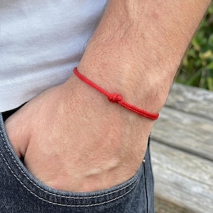Peut inclure: Un bracelet en ficelle rouge noué. Le bracelet est porté au poignet, avec une chemise blanche et un jean en arrière-plan. Le bracelet est de conception simple et minimaliste.