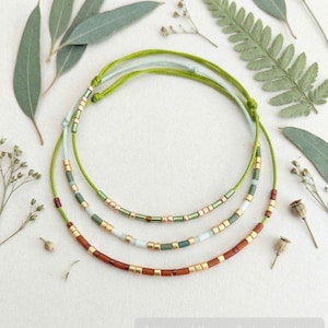 Op de afbeelding: Drie verstelbare koordarmbanden met een mix van metallic gouden, matgroene, lichtblauwe en donkerbruine cilindrische kralen. De armbanden zijn in concentrische cirkels gerangschikt, met de tekst https://braceletsrt.etsy.com zichtbaar.