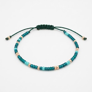 Può includere: Braccialetto regolabile con perline turchesi e dorate. Il braccialetto ha una chiusura a nodo macramè verde scuro e cordoncini verdi abbinati. Le estremità dei cordoncini hanno piccole perline dorate.