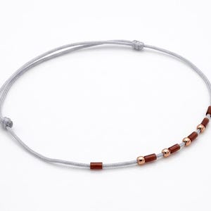 Può includere: Un braccialetto alla caviglia minimalista con un cordoncino grigio chiaro e una serie di piccole perline cilindriche marrone scuro e perline sferiche color oro rosa. Il braccialetto è annodato ad ogni estremità.