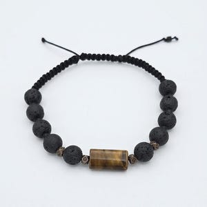 Op de afbeelding: Een zwarte lavasteen armband met een rechthoekige tijgeroogsteen in het midden. De armband heeft een verstelbaar zwart koord en kleine decoratieve kralen. De lavastenen zijn rond en hebben een textuur.