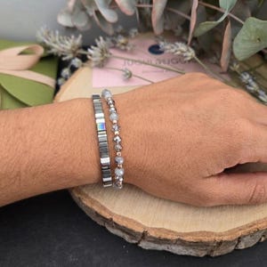 Op de afbeelding: Twee armbanden om een pols. Eén armband heeft rechthoekige, iriserende kralen en de andere heeft kleine, ronde, grijze kralen. De armbanden worden getoond op een houten schijf, met een groene geschenkdoos en gedroogde bloemen op de achtergrond.