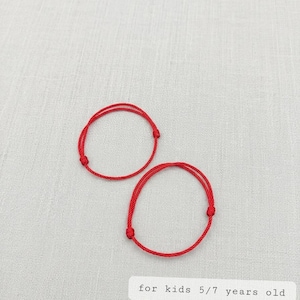Könnte beinhalten: Zwei rote Schnurarmbänder, geknotet und kreisförmig, werden auf einem hellgrauen Stoff gezeigt. Der Text "for kids 5/7 years old" ist auf einem weißen Rechteck darunter aufgedruckt. Diese Armbänder sind für Kinder.
