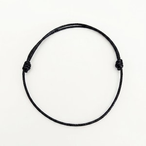 Peut inclure: Un bracelet noir réglable, fait d'un cordon fin et tressé. Le bracelet comporte deux nœuds pour ajuster la taille. Le fond est blanc uni. L'URL de la boutique Etsy est visible en bas.