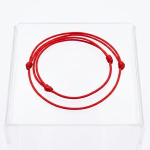 May include: A vibrant red bracelet with a knotted design. The bracelet is made of a thin, flexible material and is displayed on a clear, square stand. The design features three knots.