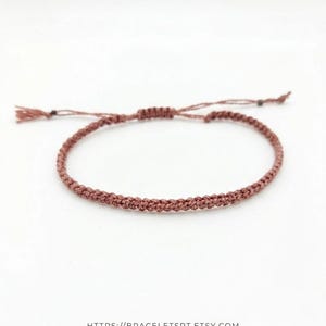 Peut inclure: Un bracelet fin tressé de couleur rose poudré doté d'un fermoir à nœud coulissant pour l'ajustement. Le cordon est tissé selon un motif serré et uniforme. Le texte HTTPS://BRACELETSRT.ETSY.COM est imprimé au bas de l'image.