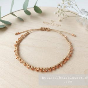 Op de afbeelding: Een beige macramé armband met kleine metallic koperkleurige kralen verwerkt in het koord. De armband heeft een verstelbare schuifknoopsluiting met twee dunne uiteinden, elk afgewerkt met een bijpassende koperkleurige kraal.