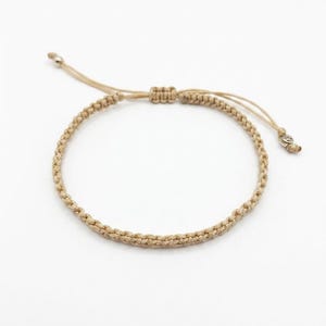 Peut inclure: Un bracelet en cordon beige au design tressé doté d'un fermoir coulissant réglable. Les extrémités du cordon sont terminées par de petites perles métalliques, offrant un ajustement sûr et flexible pour cet accessoire de poignet tissé.