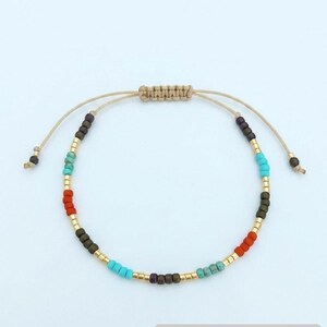 Könnte beinhalten: Ein handgefertigtes Perlenarmband mit Makramee-Verschluss. Das Armband hat eine Vielzahl farbiger Perlen, darunter Türkis, Rot, Gold und Dunkelbraun. Das verstellbare Armband ist aus beigem Kordel gefertigt und wird auf weißem Hintergrund präsentiert. Etsy-Shop-URL sichtbar.