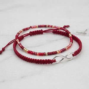 Può includere: Due braccialetti rossi. Uno è in stile macramè con un ciondolo infinito argentato. L'altro ha perline rosse, rosa e dorate. I braccialetti sono fatti a mano e regolabili.