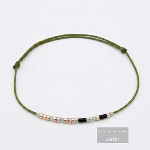 Può includere: Un braccialetto con cordino verde oliva con una serie di piccole perline. Le perline sono un mix di oro rosa, verde chiaro e nero. La parola "sister" è in un piccolo riquadro.