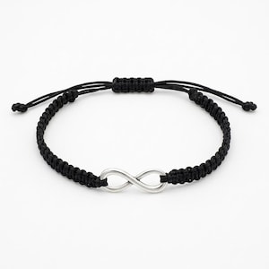 Könnte beinhalten: Schwarzes Makramee-Armband mit einem silberfarbenen Unendlichkeitszeichen. Das verstellbare Armband besteht aus gewebtem schwarzem Kordel und hat einen Schiebeknotenverschluss. Das Unendlichkeitszeichen ist ein klassisches Design, das Ewigkeit symbolisiert.