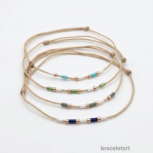 Può includere: Cinque braccialetti minimalisti con cordoncini beige e perline colorate. Ogni braccialetto presenta una combinazione di colori diversa: turchese, verde, grigio e blu navy. I braccialetti sono esposti su sfondo bianco. Il testo "braceletsrt" è visibile.