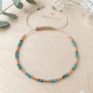Op de afbeelding: Een delicate armband met afwisselende turquoise en metallic koperkleurige kralen aan een dun beige koord. De armband is voorzien van een verstelbare schuifknoopsluiting en is gemerkt met de tekst RODICA TUGUI.