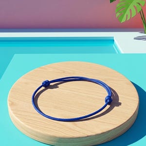 Könnte beinhalten: Ein minimalistisches blaues Armband mit verstellbaren Knoten, das auf einer hellen, kreisförmigen Holzplattform liegt. Der Hintergrund zeigt ein Pool-ähnliches Design mit einem rosa und blauen Farbverlauf und einen Papierauschnitt einer tropischen Pflanze. Die Etsy-Shop-URL ist sichtbar.