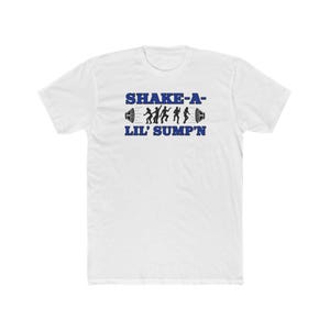May include: White t-shirt with the text "SHAKE-A-" above "LIL' SUMP'N" in blue. The design includes silhouettes of people dancing between two speaker icons. The shirt is made of a soft, comfortable material.