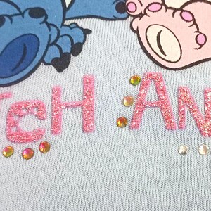 May include: Light blue fabric with the words "Stitch Angel" in pink glitter letters, embellished with rhinestones. The image features a cartoon illustration of a blue alien and a pink alien foot.