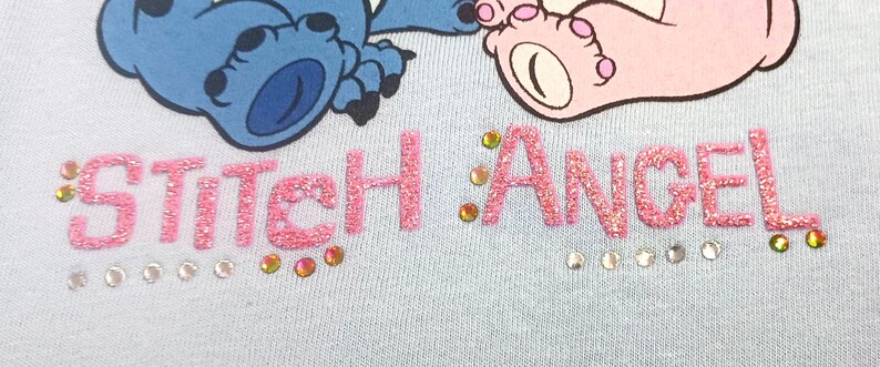 May include: Light blue fabric with the words "Stitch Angel" in pink glitter letters, embellished with rhinestones. The image features a cartoon illustration of a blue alien and a pink alien foot.
