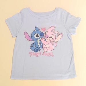 May include: A light blue t-shirt featuring a graphic of the characters Stitch and Angel embracing, with a pink heart above them. The text "Stitch Angel" is below the characters, with rhinestone accents.